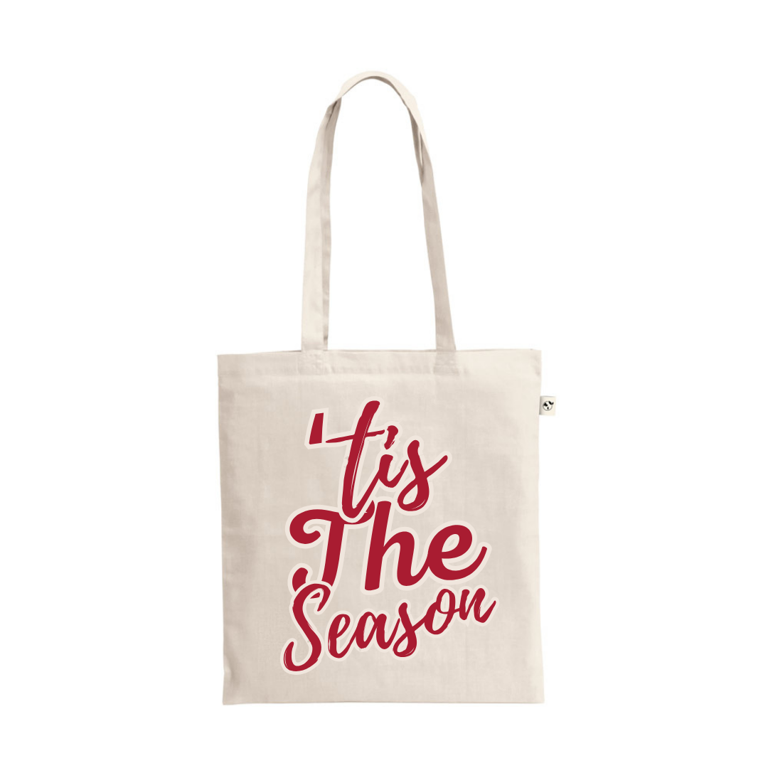 A natural cotton tote bag with the festive phrase "'Tis The Season" printed in a large, flowing, bright red script.